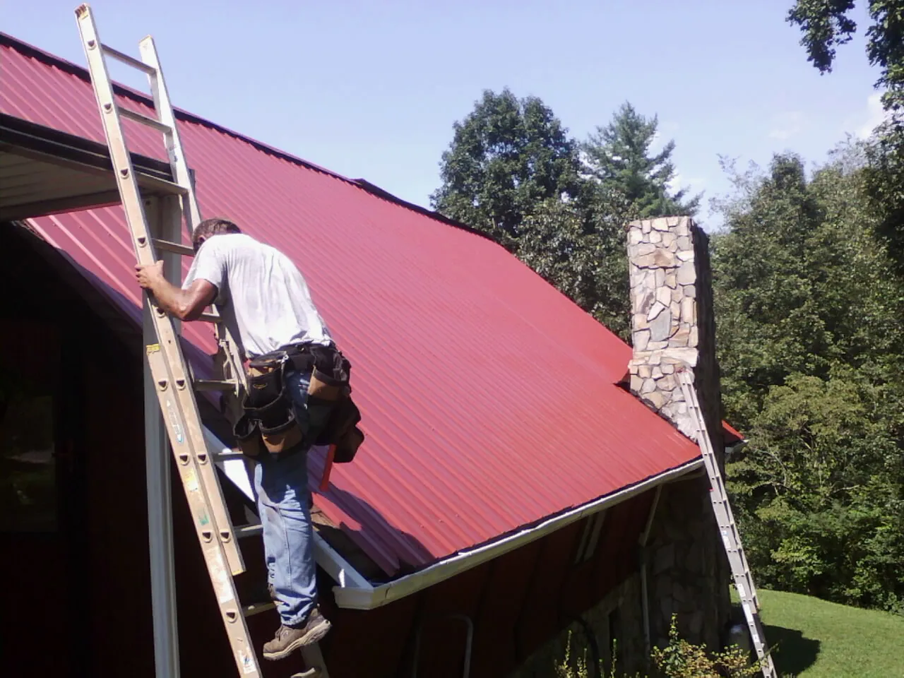 Roofer on a ladder beside a red metal roof and stone chimney — Soffit & Fascia Repair by Roofing Brothers in Cochranton